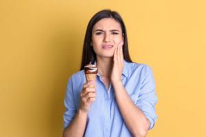 Woman holding an ice cream cone with one hand and her cheek with the other hand and a pained expression